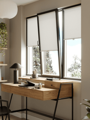This image shows white clic honeycomb blinds fitted onto a black frame tilt and turn window, above a wooden desk.