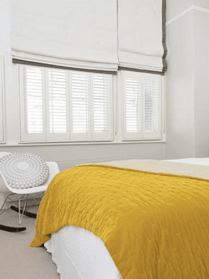 This image shows white shutters fitted onto bedroom windows, half covered by white and grey roman blinds.