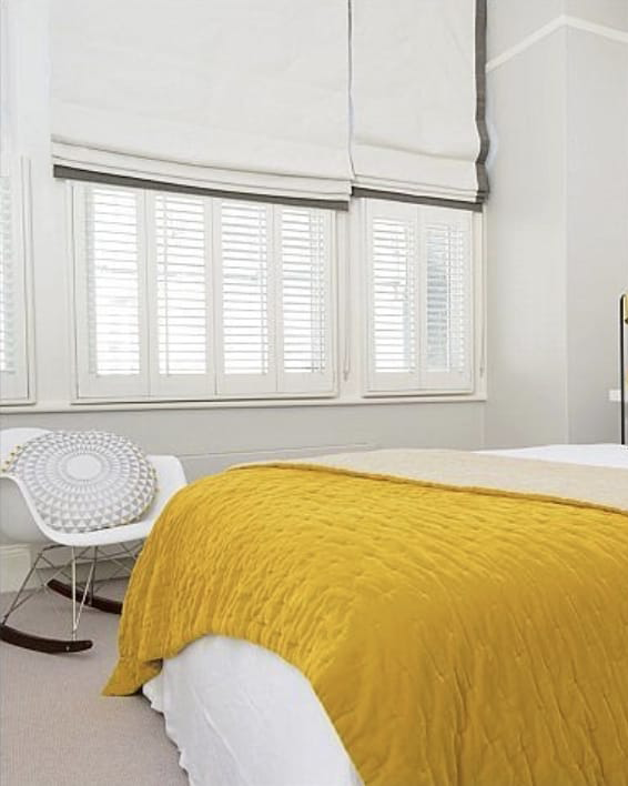 This image shows white shutters fitted onto bedroom windows, half covered by white and grey roman blinds.