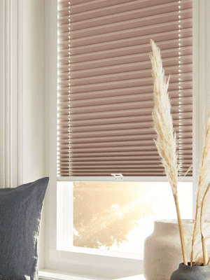 This image shows sand coloured perfect-fit honeycomb blinds fitted onto a white window frame with the sun shining through.