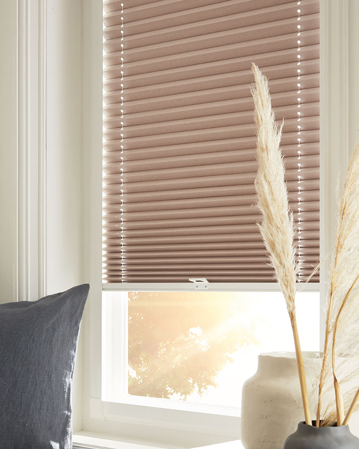 This image shows sand coloured perfect-fit honeycomb blinds fitted onto a white window frame with the sun shining through.